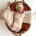 Newborn baby wrapped in beige swaddle and beanie, lying in a woven basket with brown and white blankets.